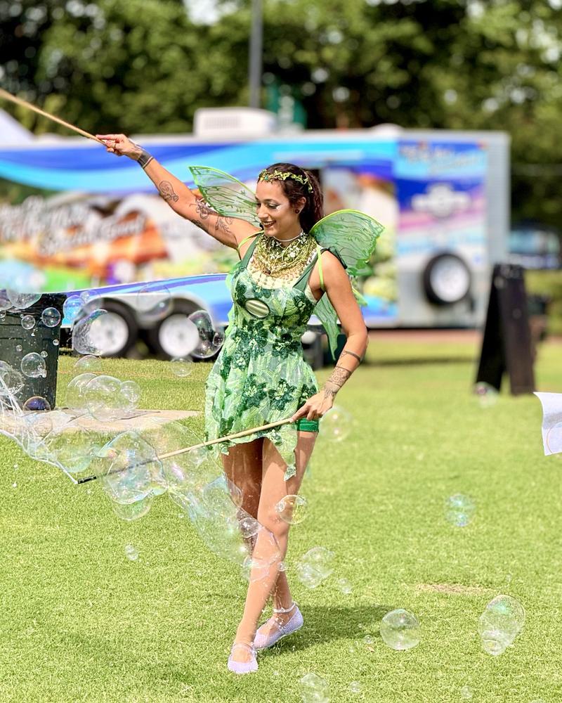 The bubble fairy in a green costume and wings, waving a bubble wand and trailing bubbles across the grass