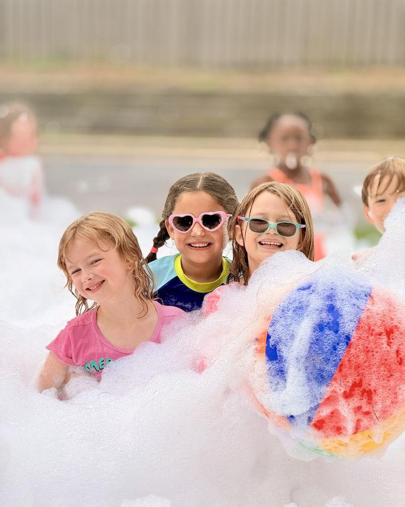 Three laughing kids in sunglasses surrounded by foam, with a foam-covered beach ball in front