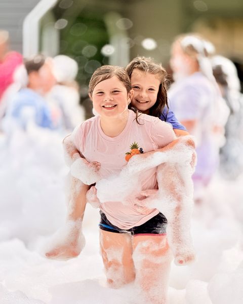 Two grinning girls covered in foam, one giving the other a piggyback ride