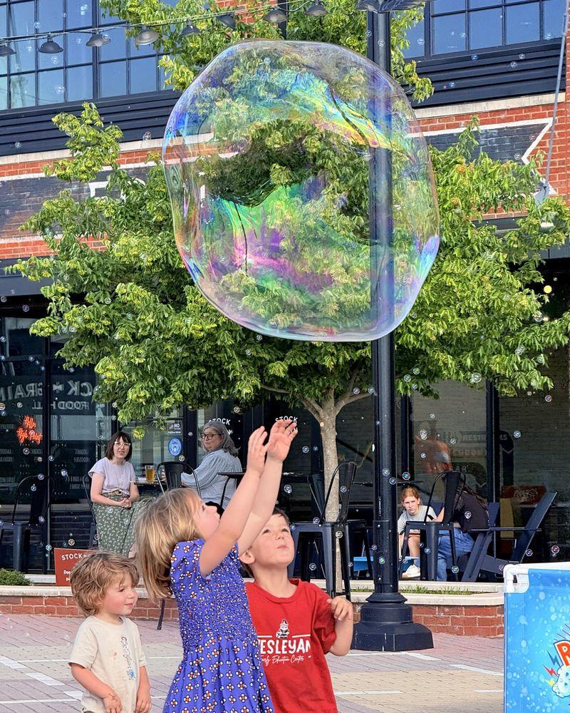 Three young children reaching up toward a giant rainbow-sheened soap bubble on a brick plaza