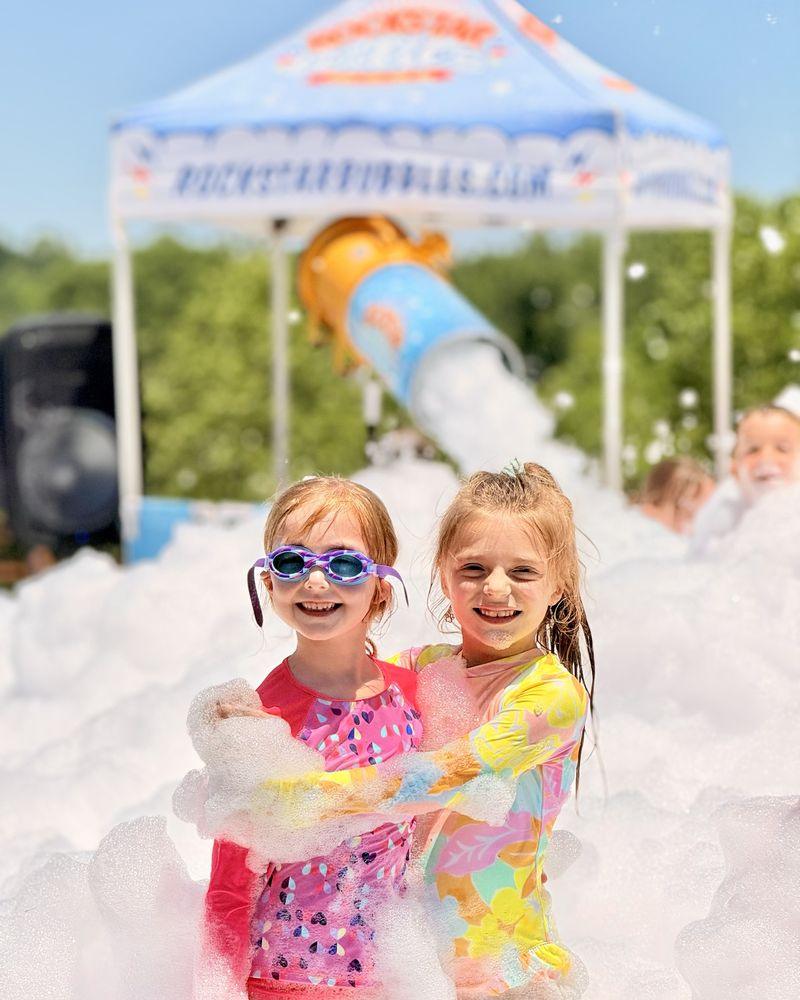 Two smiling girls hugging, waist-deep in foam, with the Rockstar Bubbles foam cannon and tent behind them